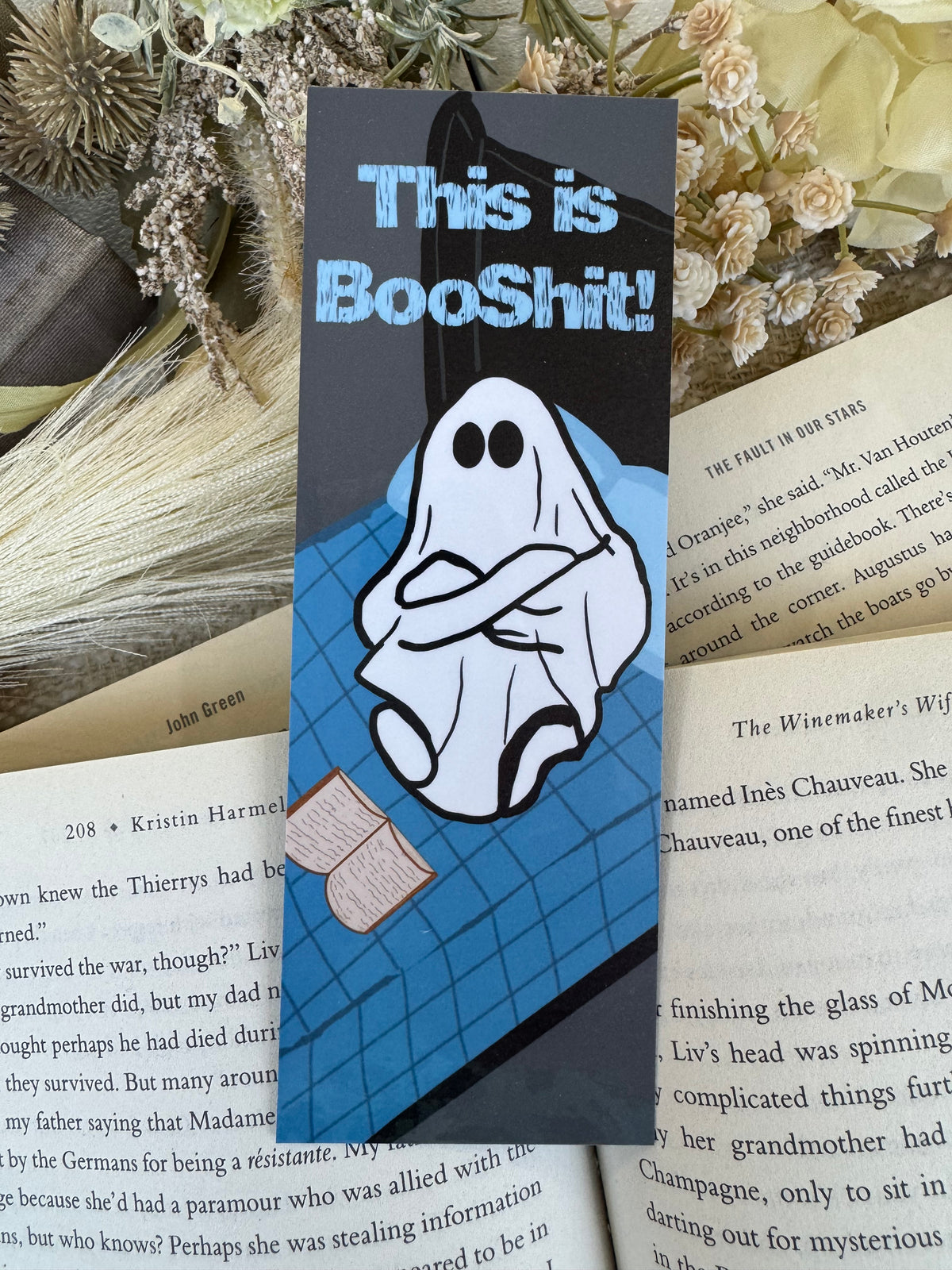 A rectangular black book marker with a cartoon ghost and the text 'This is BooShit!' lies on top of open book pages, with dried flowers in the background.