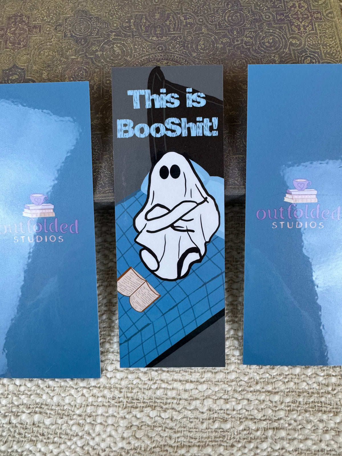Three printed bookmarks showing the front and back design of the bookmark. The bookmark features a frustrated looking ghost sitting on a bed with his book open and his arms crossed. The top says "this is booshit!"