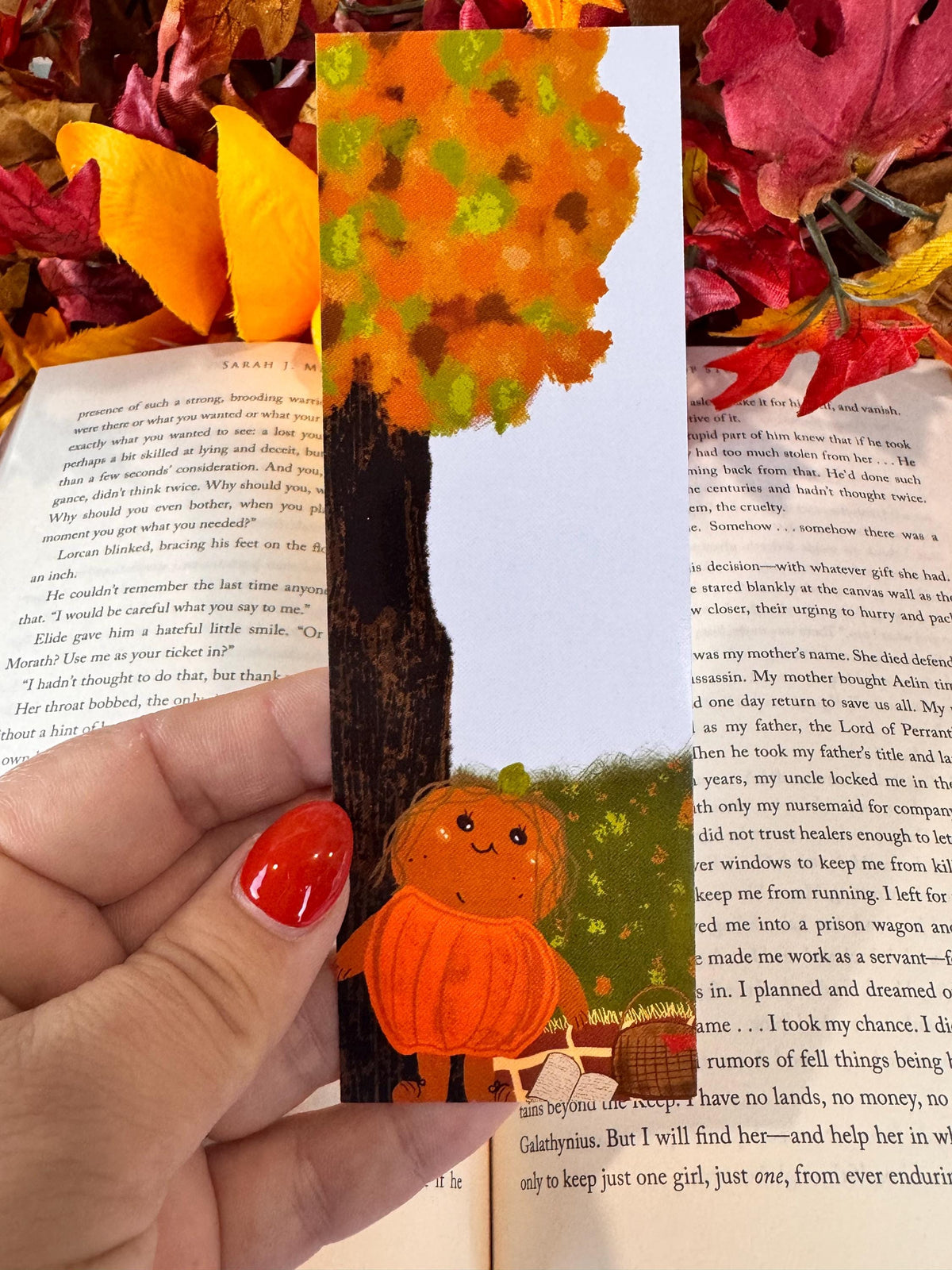Patch the pumpkin bookmark beneath an orange autumn tree, held over an open book