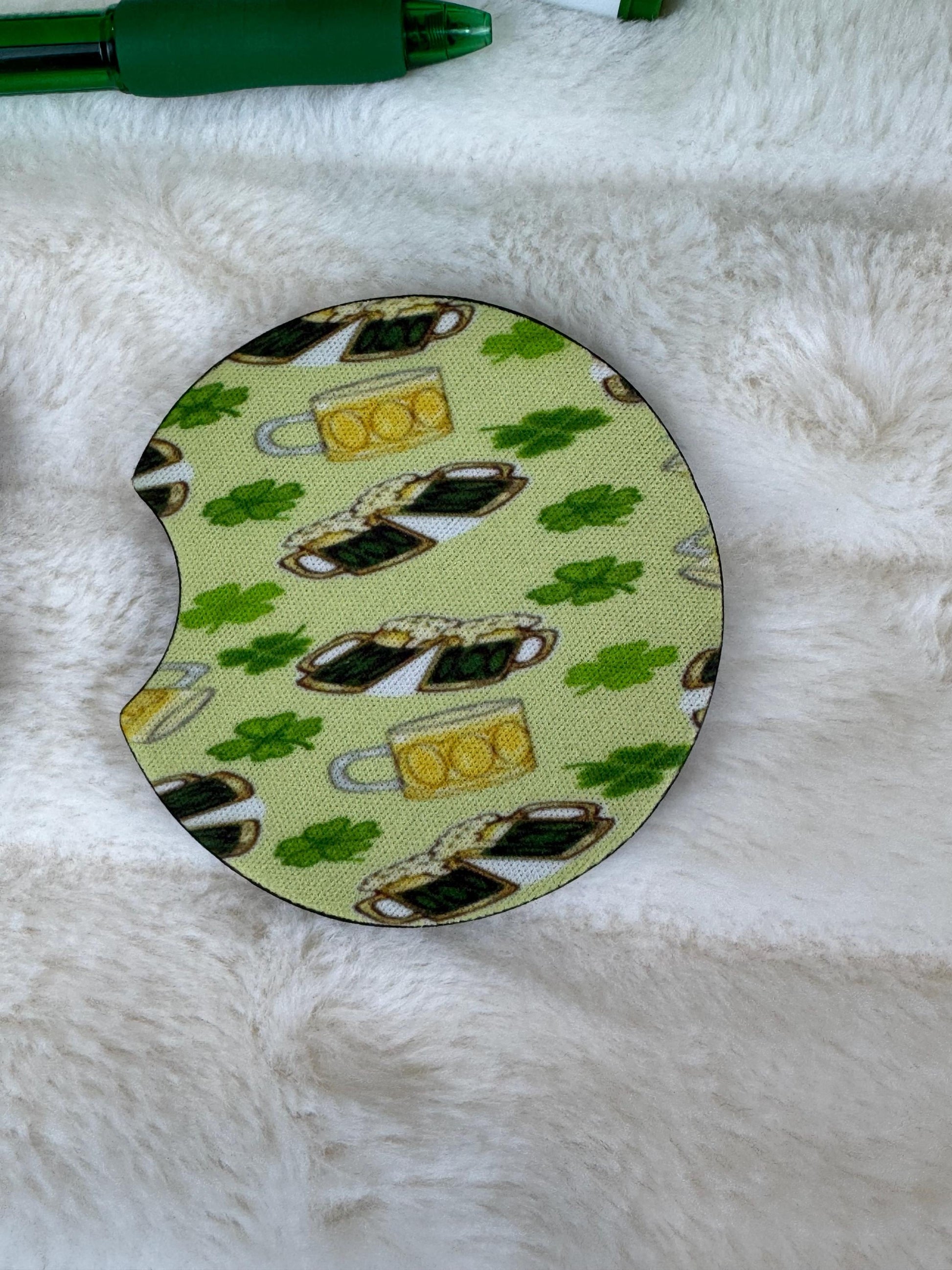 A circular car coaster with a St. Patrick's Day theme, featuring a green shamrock and beer mug pattern on a pale yellow background, placed on a white faux fur surface.