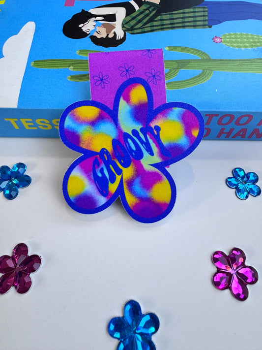 A flower-shaped magnetic bookmark with a tie-dye pattern in purple, blue, and yellow, featuring the word 'Groovy' in blue script. The bookmark is displayed on a white surface with decorative flower-shaped stickers and a colorful book in the background.
