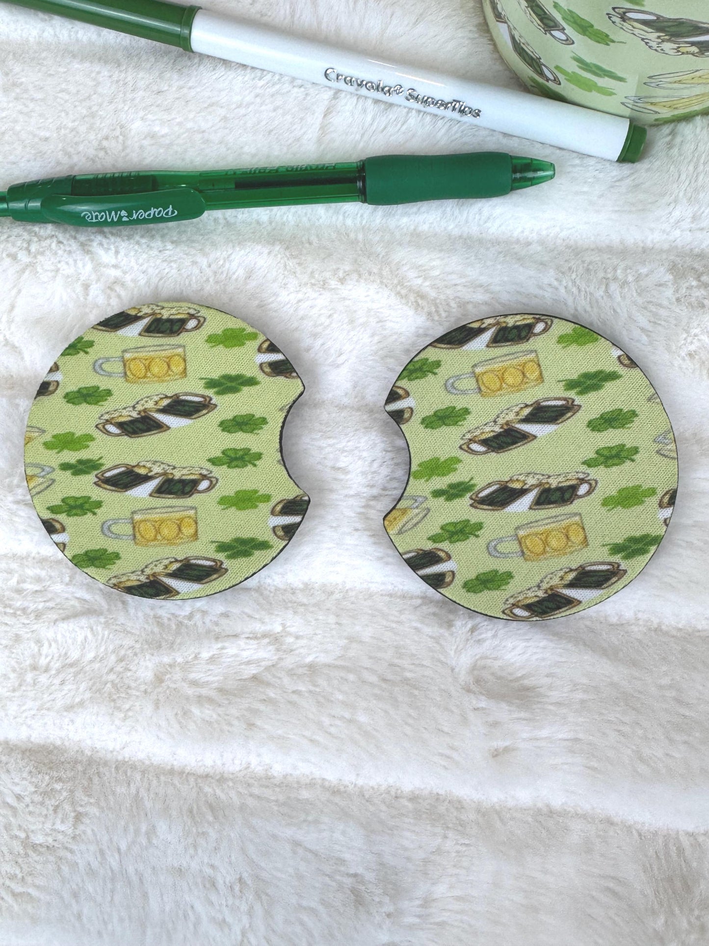 Two circular coasters with a St. Patrick's Day theme, featuring a green and beige pattern of shamrocks, leprechauns, and beer mugs, displayed on a white faux fur surface next to a green pen.