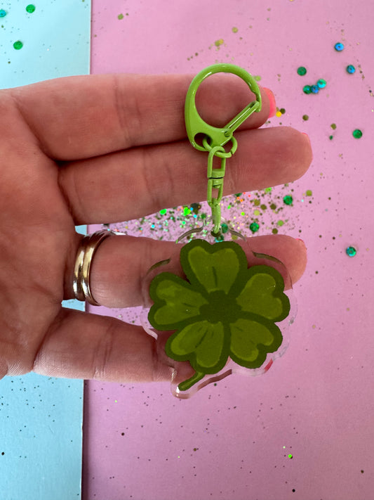 Hand holding a green four-leaf clover-shaped clip against a pink and blue background with glitter.