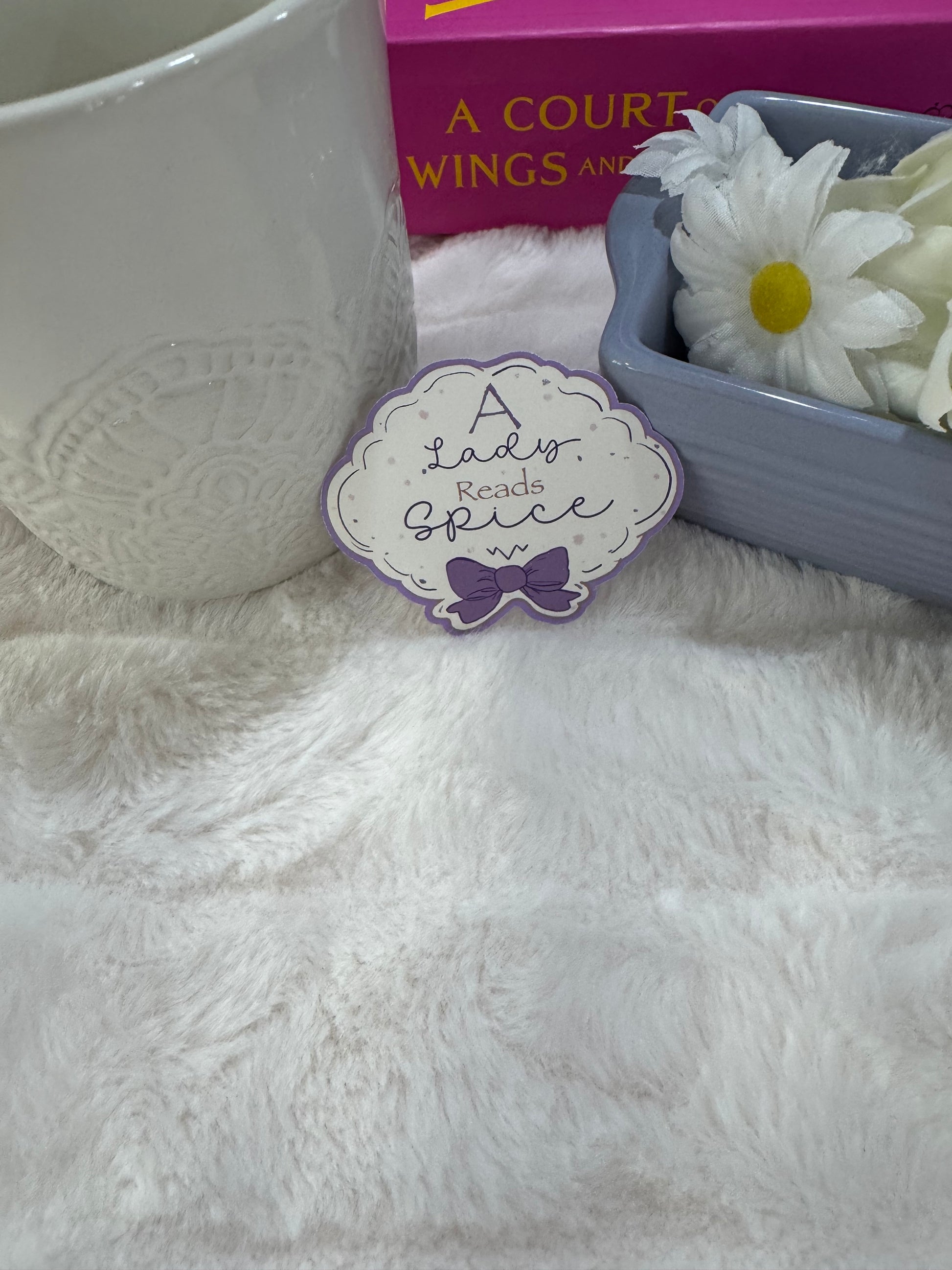 A decorative sticker or label reading 'A Lady Reads Spice' with a purple bow, placed on a white faux-fur surface next to a white ceramic mug, a pink book, and a small dish with white flowers.