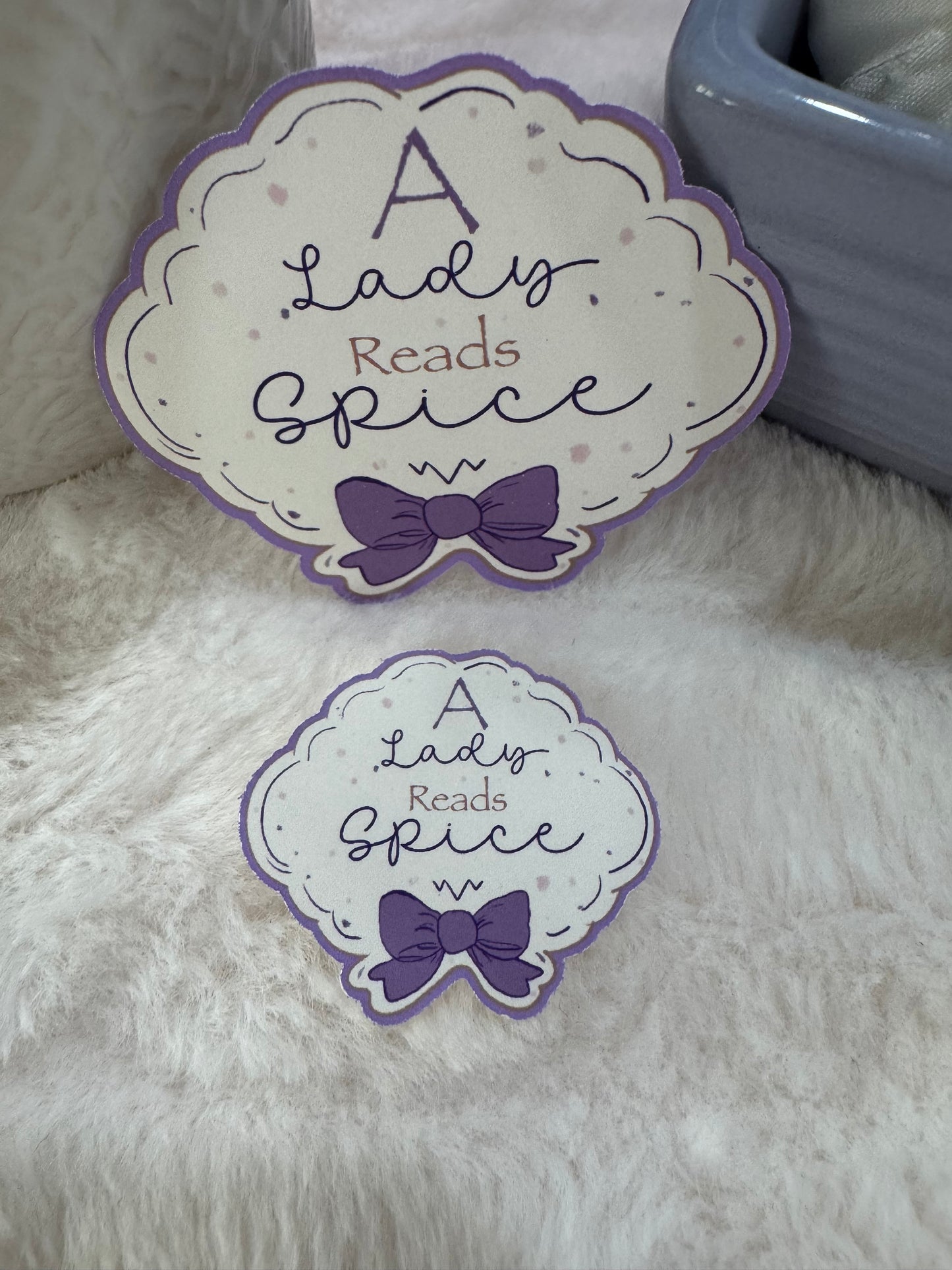 Two decorative die-cut stickers with the phrase 'A Lady Reads Spice' and a purple bow, displayed on a white faux fur surface next to a gray ceramic container.