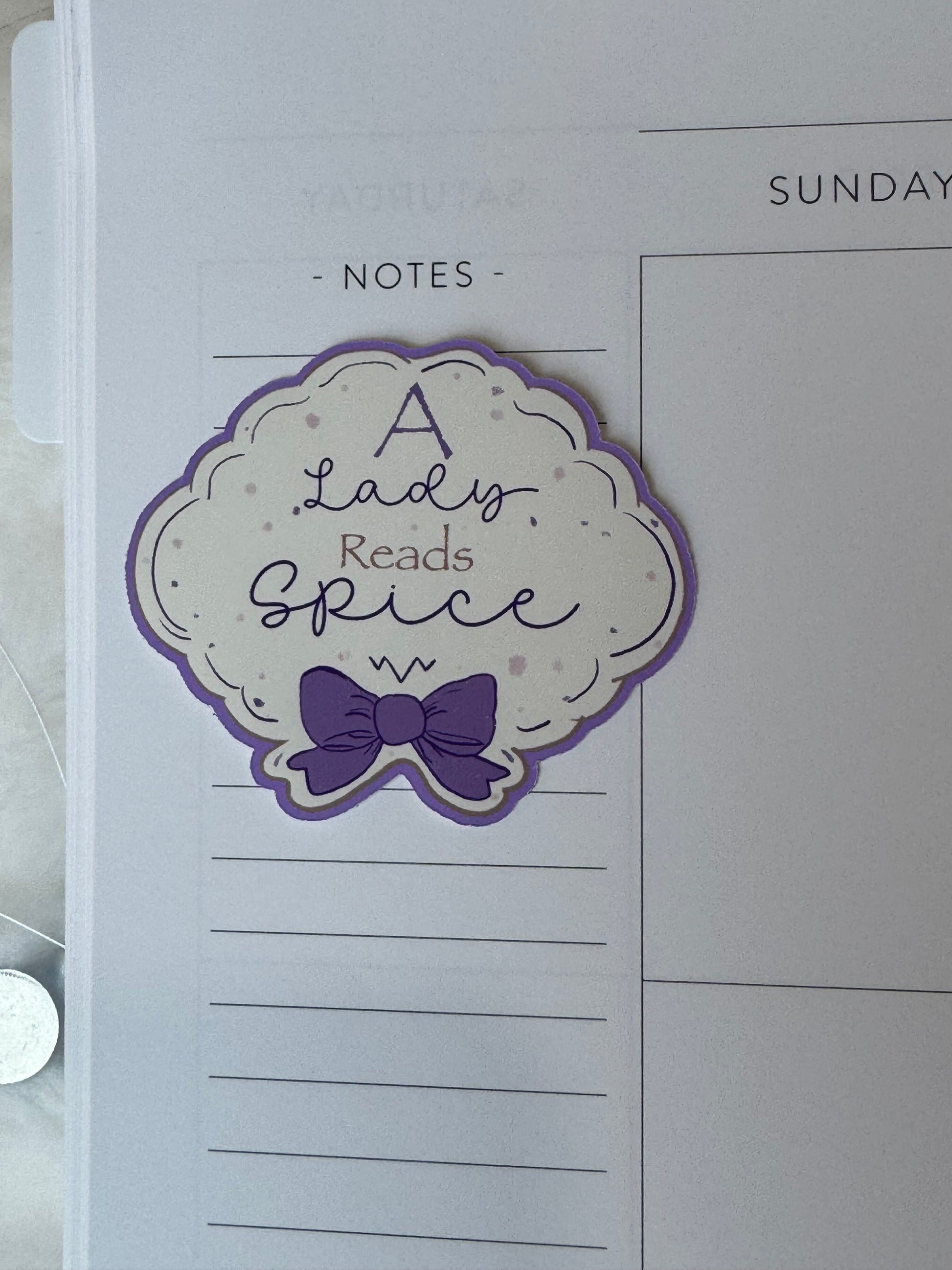 A decorative planner sticker with the phrase 'A Lady Reads Spice' in purple and peach script, featuring a purple bow and scalloped cloud border on a cream background, shown on a white lined notes page.