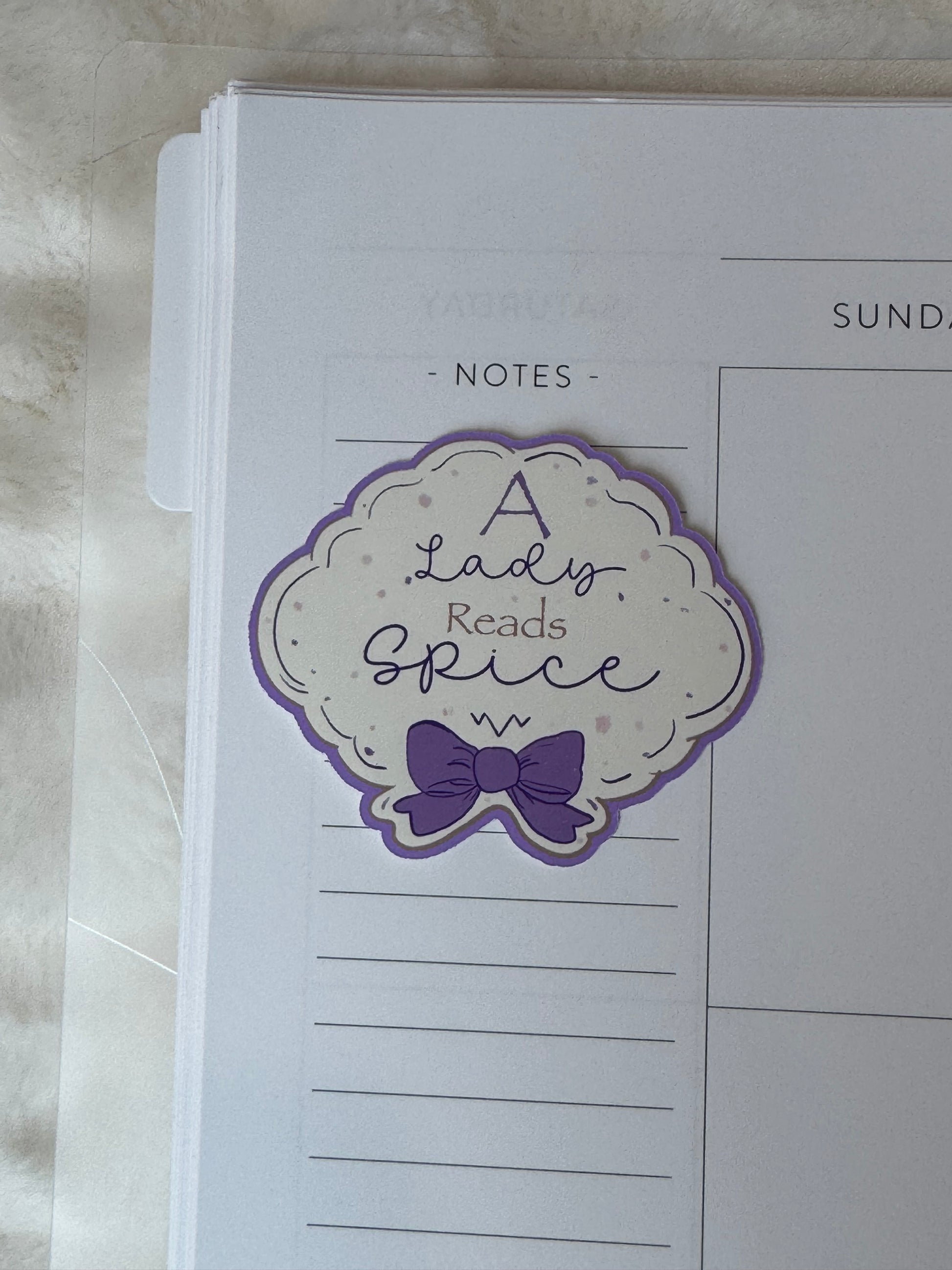 A die-cut planner sticker with the phrase 'A Lady Reads Spice' in purple script and a purple bow illustration, shown placed on a white lined notes page.