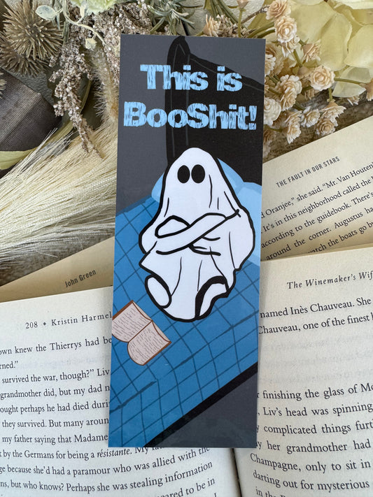 A rectangular black book marker with a cartoon ghost and the text 'This is BooShit!' lies on top of open book pages, with dried flowers in the background.