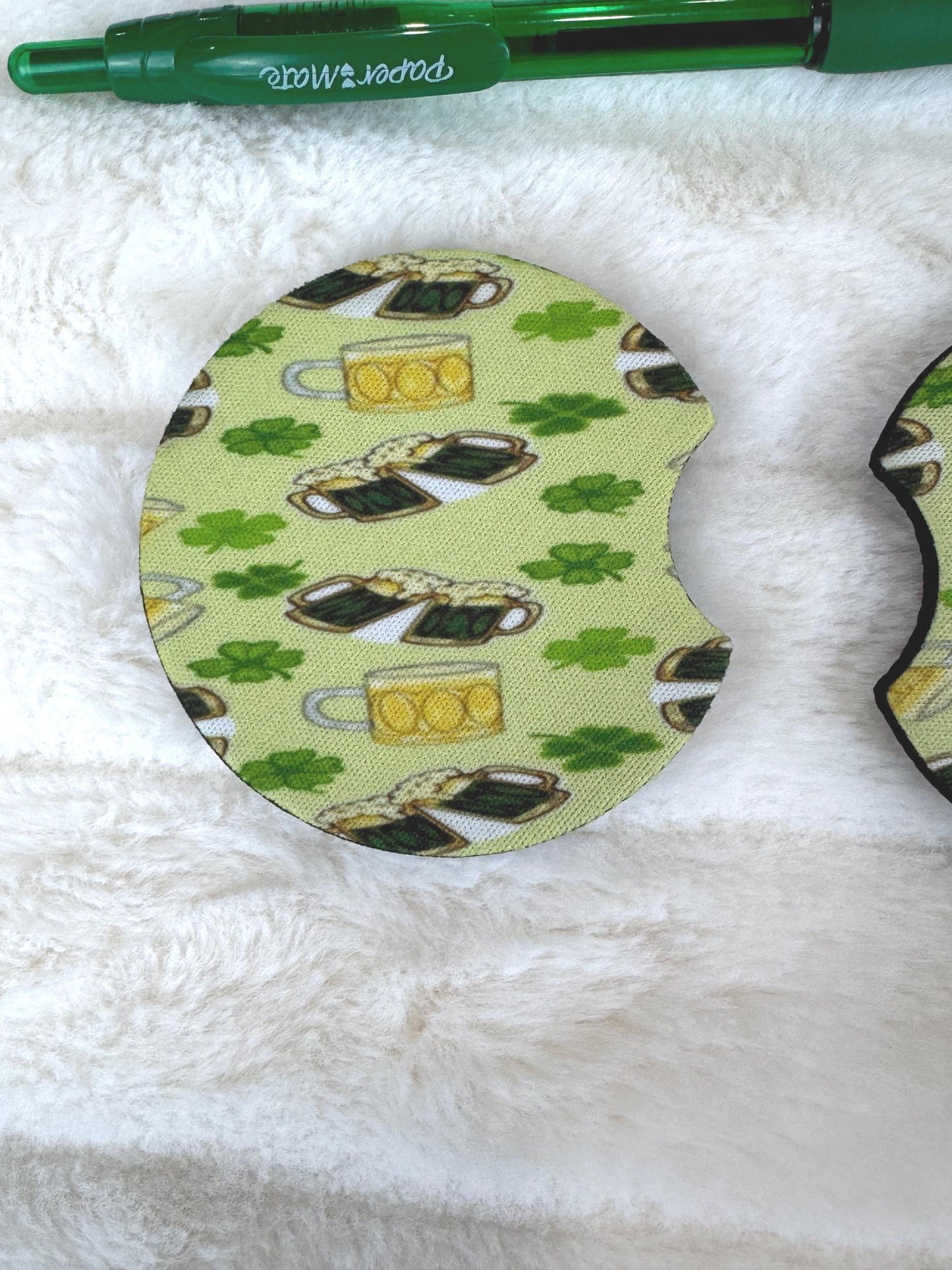 A circular St. Patrick's Day coaster with a green shamrock and beer mug pattern, displayed on a white faux fur surface next to a green pen.