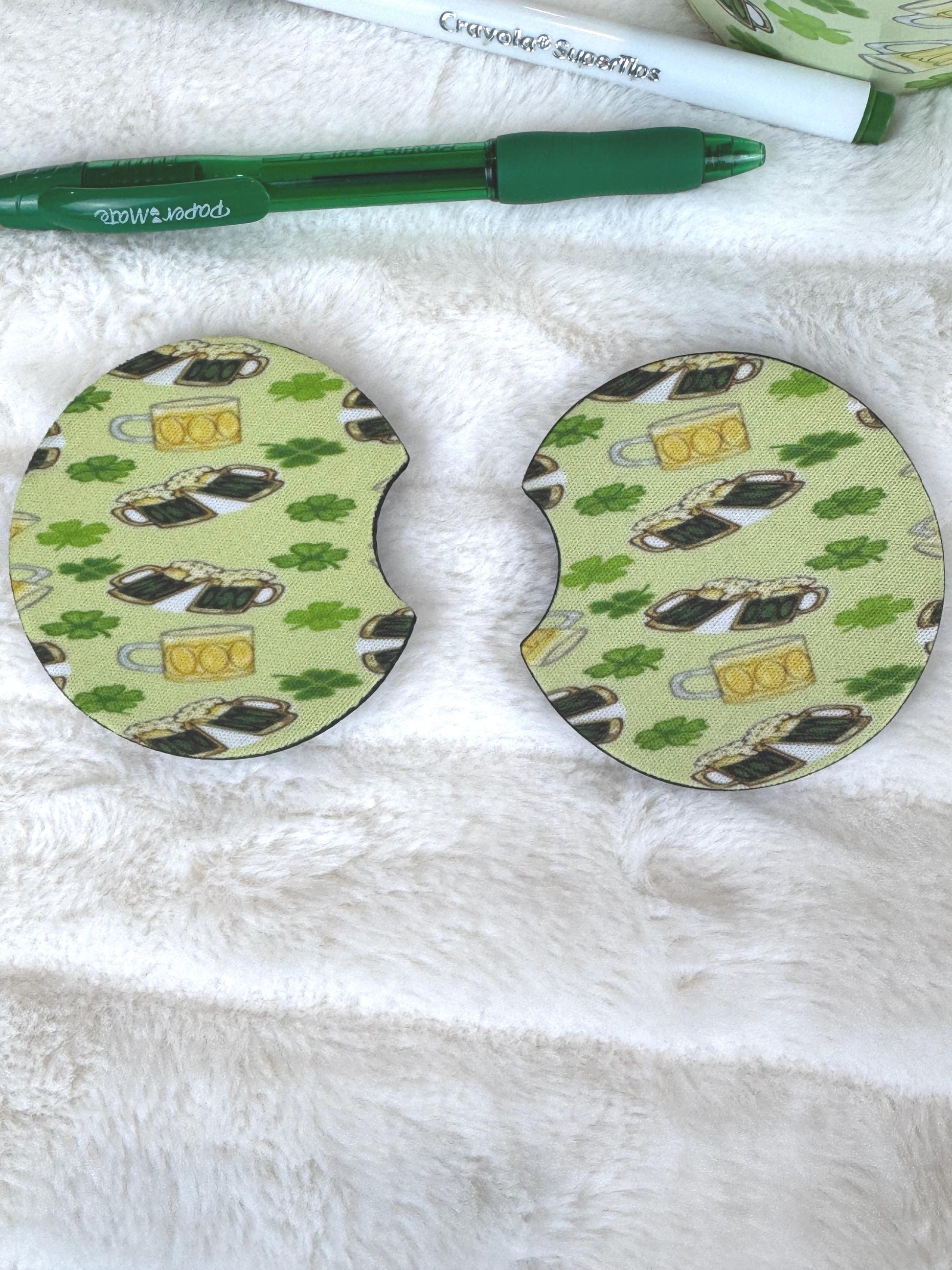 Two oval-shaped coasters with a St. Patrick's Day theme, featuring a green background with shamrocks, beer mugs, and clovers, displayed on a white faux fur surface next to a green pen.