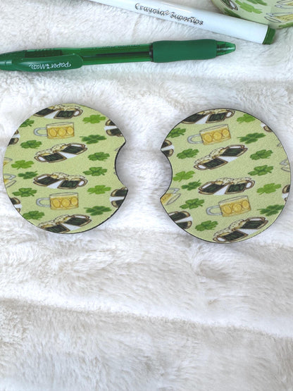 Two oval-shaped coasters with a St. Patrick's Day theme, featuring a green background with shamrocks, beer mugs, and clovers, displayed on a white faux fur surface next to a green pen.