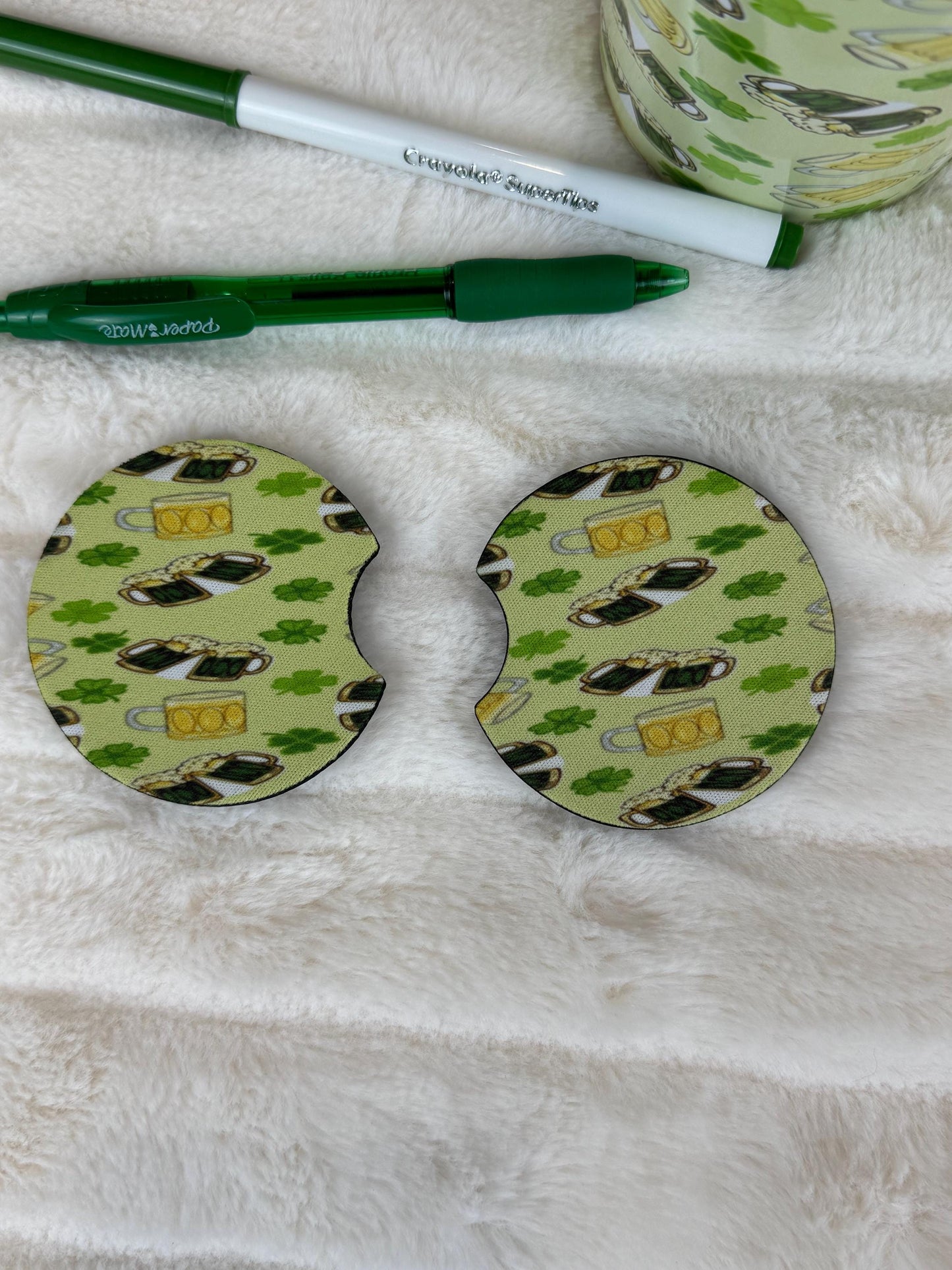 Two round coasters with a St. Patrick's Day coffee cup and clover pattern, displayed on a white faux fur surface next to a green pen and a matching patterned mug.