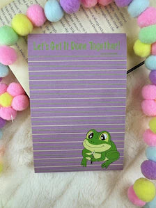 A purple striped notepad with a green cartoon frog and the text 'Let's Get It Done Together!' at the top, placed on a white furry surface with colorful pom-pom garlands around it.